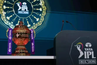 एक आईपीएल नीलामी का दृश्य जिसमें नीलामीकर्ता (ऑक्शनर) मंच पर हथौड़ा (गैवल) लिए खड़े हैं और नीचे बैठी फ्रेंचाइजी टीमों के मालिक बोली लगा रहे हैं। पृष्ठभूमि में बड़ी स्क्रीन पर खिलाड़ी की कीमत दिख रही है।