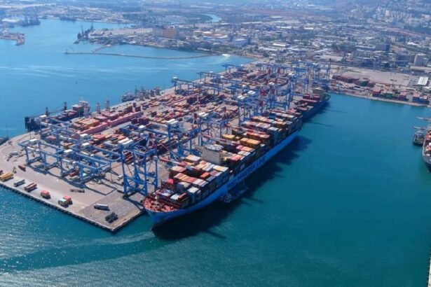 इजरायल के हाइफा बंदरगाह का हवाई चित्र, जहाज लंगर डाले, कंटेनर स्टैक, समुद्र तट पर। अडानी लोगो दिखे।