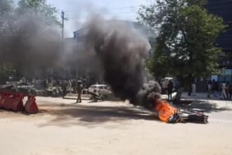 नोएडा में श्रमिक हिंसा मामले की जांच से जुड़ी प्रतीकात्मक तस्वीर, जिसमें पुलिस अधिकारी दस्तावेजों की जांच करते दिखाई दे रहे हैं और पृष्ठभूमि में प्रदर्शन का दृश्य नजर आ रहा है