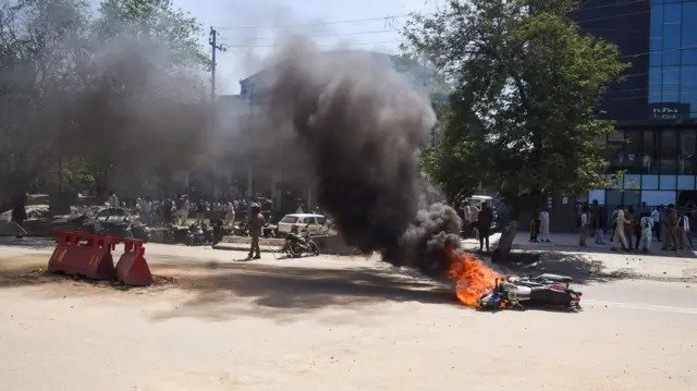 नोएडा में श्रमिक हिंसा मामले की जांच से जुड़ी प्रतीकात्मक तस्वीर, जिसमें पुलिस अधिकारी दस्तावेजों की जांच करते दिखाई दे रहे हैं और पृष्ठभूमि में प्रदर्शन का दृश्य नजर आ रहा है