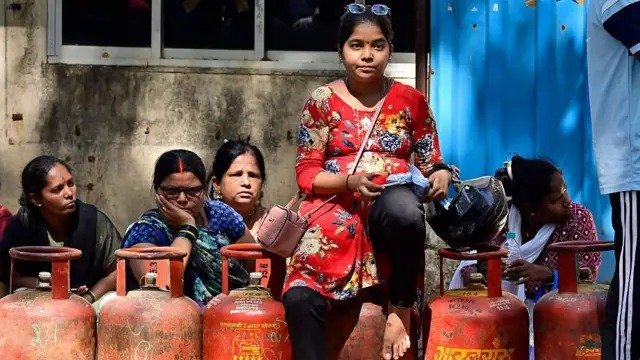 घरेलू LPG गैस सिलेंडर की तस्वीर जिसमें बढ़ती कीमतों का ग्राफिक ओवरले दिखाया गया है और पीछे रसोई का दृश्य धुंधला दिखाई दे रहा है